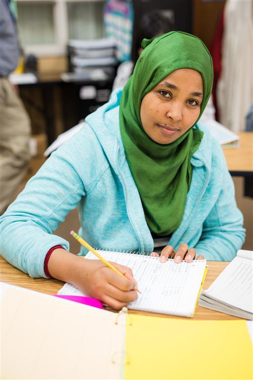 Student writing on paper and looking at camera. 